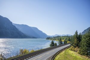 A railroad track next to a highway road with lane dividing opposite traffic runs along the scenic Columbia River bank with green trees and rocky mountains in Oregon